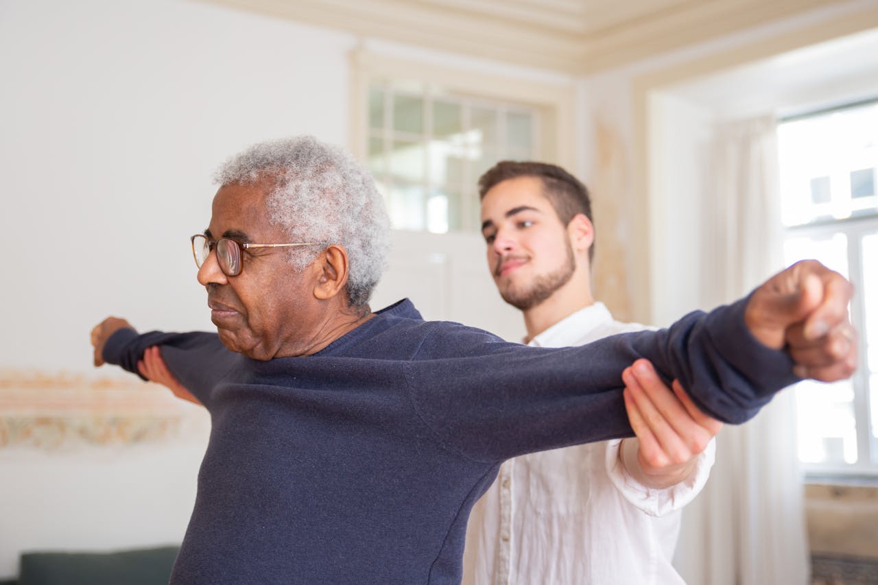 journey A senior man is assisted by a caregiver for stretching exercises indoors.
